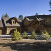 The Cedar Style by Schiffer Publishing, Ltd. is a large two-story log cabin with multiple gable roofs, a three-car garage, covered front porch, and stone driveway edged with wood chips and shrubs amidst pine trees.