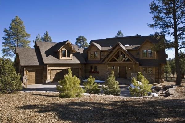 The Cedar Style by Schiffer Publishing, Ltd. is a large two-story log cabin with multiple gable roofs, a three-car garage, covered front porch, and stone driveway edged with wood chips and shrubs amidst pine trees.