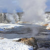 Experience Yellowstone National Park with Schiffer Publishing, Ltd.: snow blankets the landscape as a river winds past steaming geysers and snow-covered trees beneath a clear sky.