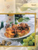 A plate of roasted quail, featured in "Cooking Wild Game" by Schiffer Publishing, Ltd., rests on vegetables and herbs, surrounded by green grapes, quail eggs, a wine glass, and a bottle atop a white tablecloth.