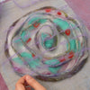 A hand arranges purple, turquoise, white, and red wool fibers in a spiral on a mesh mat, using classic felting techniques. Featured: Felting by Schiffer Publishing, Ltd.