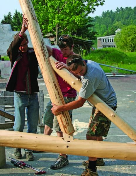 Workers assemble large wooden logs outdoors using sustainable techniques from "Natural Building" by Schiffer Publishing, Ltd. Trees and a building are in the background, with tools and wood shavings scattered nearby.