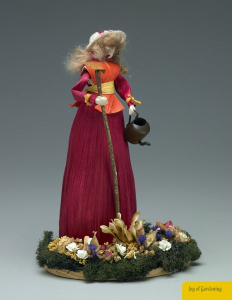Discover Appalachian craft with "Making Colorful Corn Shuck Dolls" by Schiffer Publishing, Ltd.—a guide inspired by figurines like the woman in a red dress and bonnet, holding a watering can with "Joy of Gardening" on a base of artificial flowers.