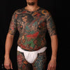 A man in a white loincloth stands against a black background, his body adorned with colorful Japanese Style Tattoo Art by Schiffer Publishing, Ltd., featuring intricate figures, flowers, and designs.