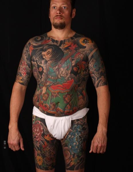 A man in a white loincloth stands against a black background, his body adorned with colorful Japanese Style Tattoo Art by Schiffer Publishing, Ltd., featuring intricate figures, flowers, and designs.