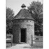 A round brick dovecote with a conical shingled roof stands amid grass and trees, ringed by a wooden fence. Its open doorway recalls the eerie stories found in "Haunted Plantations of Virginia" from Schiffer Publishing, Ltd.