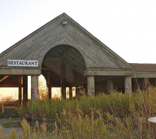 “Gone But Not Forgotten” by Schiffer Publishing, Ltd. captures the haunting allure of an abandoned restaurant, sunlight streaming through its open walls as overgrown weeds and faded signs reflect the lost charm of New England ghost towns.