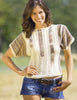 A smiling woman with wavy brown hair wears "Patchwork Knitting" by Schiffer Publishing, Ltd.—a short-sleeved, crocheted beige and brown top—paired with denim shorts, standing outdoors with one hand on her hip.