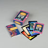The Audacious Action Angels Oracle features a colorful deck of cards, a matching pink box, and an illustrated booklet, all arranged on a light gray surface. Some oracle cards are spread out, showcasing their vibrant designs and images.
