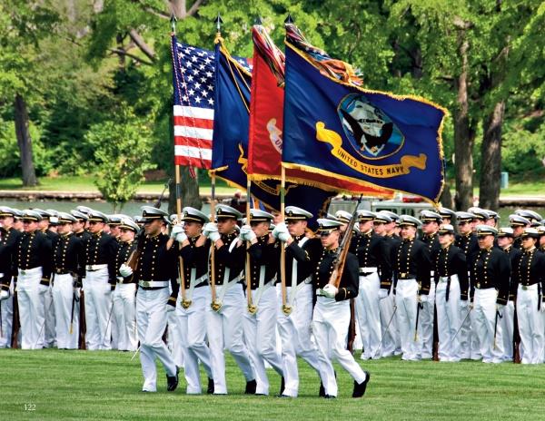 A group of U.S. Naval Academy officers in dress uniform march with the American flag on a grassy field in Annapolis, as depicted in "Annapolis Reflections" by Schiffer Publishing, Ltd.