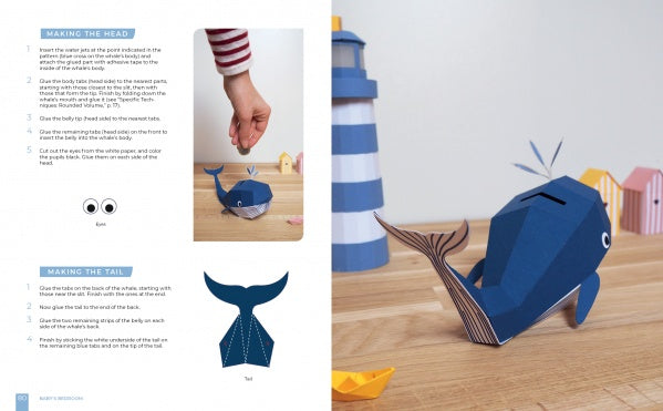 The "Paper Joy for Every Room" guide by Schiffer Publishing, Ltd. features step-by-step photo instructions to assemble a blue paper whale craft, shown finished on a wooden table beside a lighthouse and decorative houses.
