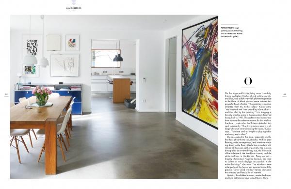 A modern, minimalist dining and living area inspired by German design—featuring details from "Home Story" by Schiffer Publishing, Ltd.—has white walls, abstract art, a wooden table with flowers, a blue chair, an open kitchen, and abundant natural light.