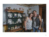 A woman and a man stand together, smiling, in a cozy room filled with mementos. Their commemorative tattoos, inspired by "War Paint" from Schiffer Publishing, Ltd., hint at veterans' stories and personal histories.