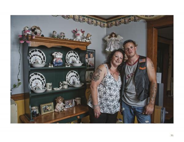 A woman and a man stand together, smiling, in a cozy room filled with mementos. Their commemorative tattoos, inspired by "War Paint" from Schiffer Publishing, Ltd., hint at veterans' stories and personal histories.