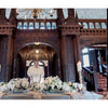 A bride in a white gown poses behind a candlelit table with white flowers in an ornate wooden hall, echoing the elegant inspiration found in *The White Dress in Color: Wedding Inspirations for the Modern Bride* by Schiffer Publishing, Ltd.