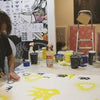 A person in a studio uses 2Create by Schiffer Publishing, Ltd., channeling street art on a large canvas with yellow and black designs amid paint supplies and artworks—a creative scene recalling New York City graffiti artists.