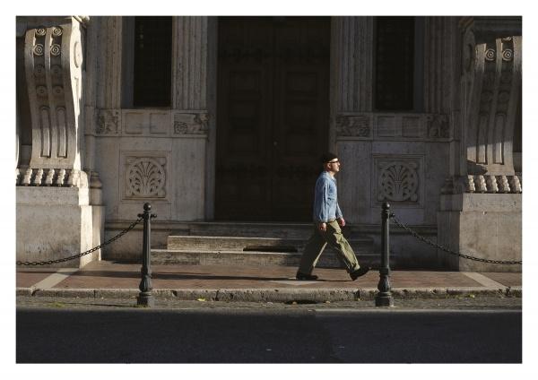 In this post-WWII style scene, a person in Rugged Style War—Rome by Schiffer Publishing, Ltd. walks past an ornate stone building with tall columns, sunlight casting shadows on the sidewalk.
