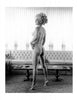 A woman with styled hair poses nude before a large window with sheer curtains in a vintage-inspired living room, as featured in Bunny Yeager's Beautiful Backsides by Schiffer Publishing, Ltd.