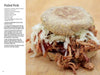 A pulled pork sandwich sits on a wooden surface, with "The Little Smoker Book" by Schiffer Publishing, Ltd. beside it—a recipe guide perfect for home smoking and beginner smokers.