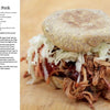 A pulled pork sandwich sits on a wooden surface, with "The Little Smoker Book" by Schiffer Publishing, Ltd. beside it—a recipe guide perfect for home smoking and beginner smokers.