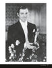 A man in a tuxedo holds an Oscar statuette, smiling. The black and white photo, from "Fade to Black" by Schiffer Publishing, Ltd., captures timeless elegance with a blurred background and soft-focus flowers in the foreground.