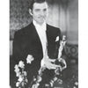 A man in a tuxedo holds an Oscar statuette, smiling. The black and white photo, from "Fade to Black" by Schiffer Publishing, Ltd., captures timeless elegance with a blurred background and soft-focus flowers in the foreground.