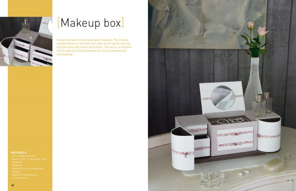 The "Vintage Cardboard Crafting" box by Schiffer Publishing, Ltd. sits on a white table beside a vase with a pink flower, a round mirror, and glass containers, against a dark gray wall. Text on the left reads Vintage Cardboard Crafting.