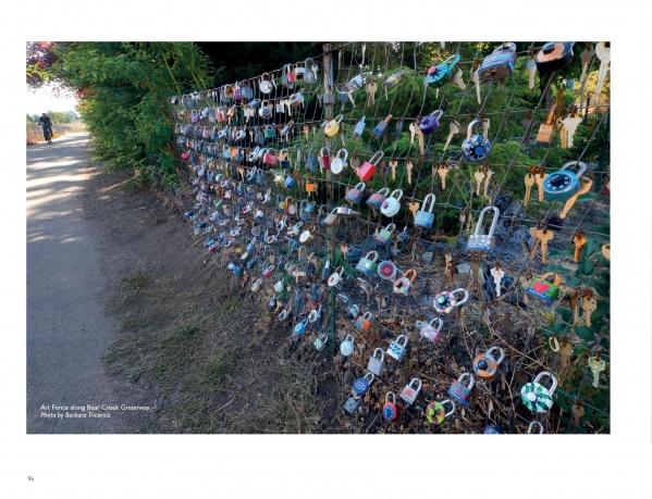 In Ashland, Oregon, a wire fence along a park path is adorned with colorful padlocks and tags, symbolizing love or memories, as described in "Ashland, Oregon" by Schiffer Publishing, Ltd. Trees and greenery frame the scenic walkway.