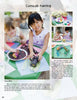 Two children sit outside, grinning as they paint corn cobs at a table filled with art supplies, inspired by activities from "Kinder Gardens" by Schiffer Publishing, Ltd. Painted corn and leaves complete this fun gardening scene.