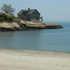 A gray house perches on rocky cliffs above a tranquil blue ocean along a scenic route from *Connecticut by Bicycle* by Schiffer Publishing, Ltd. A sandy beach and clear sky complete this peaceful coastal scene.