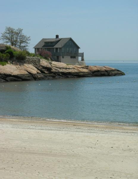 A gray house perches on rocky cliffs above a tranquil blue ocean along a scenic route from *Connecticut by Bicycle* by Schiffer Publishing, Ltd. A sandy beach and clear sky complete this peaceful coastal scene.