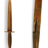 A bronze-colored Fairbairn-Sykes Fighting Knife by Schiffer Publishing, Ltd. with a ribbed handle lies beside its brown leather sheath, which has a loop and partially holds the knife on a white background.