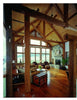 Enjoy a spacious rustic living room featuring Timber Frame Hybrids by Schiffer Publishing, Ltd., with exposed timber construction, wooden beams, large windows with water views, cozy armchairs, stone fireplace, wood floors, and a vintage trunk coffee table.