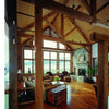 Enjoy a spacious rustic living room featuring Timber Frame Hybrids by Schiffer Publishing, Ltd., with exposed timber construction, wooden beams, large windows with water views, cozy armchairs, stone fireplace, wood floors, and a vintage trunk coffee table.