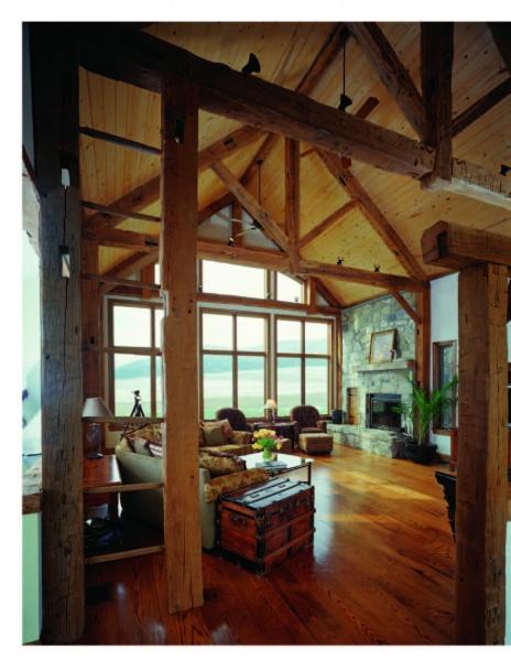 Enjoy a spacious rustic living room featuring Timber Frame Hybrids by Schiffer Publishing, Ltd., with exposed timber construction, wooden beams, large windows with water views, cozy armchairs, stone fireplace, wood floors, and a vintage trunk coffee table.