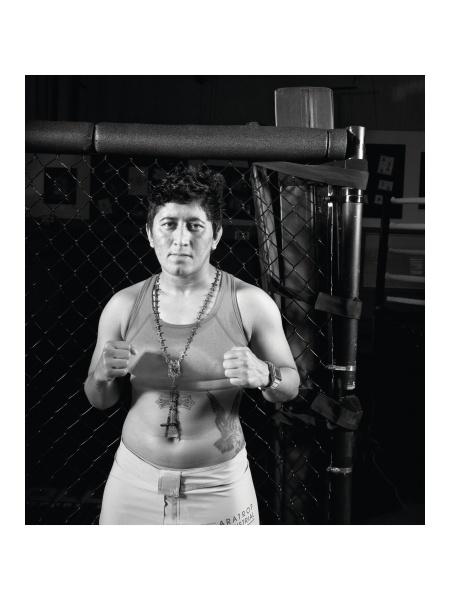 A person with short hair and a cross necklace stands in front of a chain-link fence, embodying the fierce spirit of classic fighter portraits, as featured in "Tough Guys" by Schiffer Publishing, Ltd.