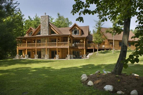 The Cedar Style by Schiffer Publishing, Ltd. features a classic log cabin with a stone chimney and wraparound porch, set on a grassy lawn with trees, landscaped rocks, and a mulch bed in the foreground.