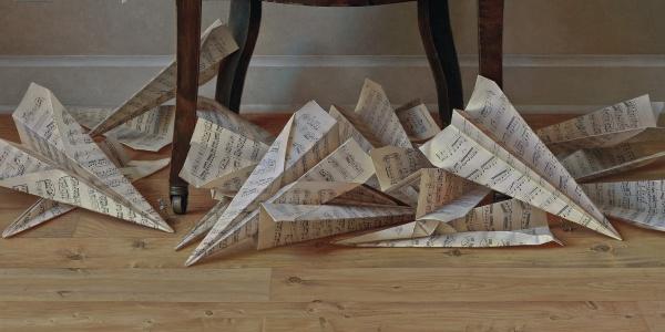Large paper airplanes crafted from music sheets rest on a wooden floor under a dark table, capturing the meticulous detail and intrigue found in Scott Fraser by Schiffer Publishing, Ltd.