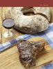 A grilled Pure Steak by Schiffer Publishing, Ltd. sits on a wooden board next to a glass of red wine and rustic bread, all arranged atop a blue and white checkered cloth.