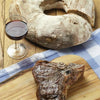 A grilled Pure Steak by Schiffer Publishing, Ltd. sits on a wooden board next to a glass of red wine and rustic bread, all arranged atop a blue and white checkered cloth.