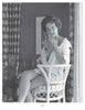 A woman sits on a wicker chair in classic 1950s style, smoking and gazing at the camera. This black-and-white image captures the allure featured in "Femmes Fatales of the 1950s" by Schiffer Publishing, Ltd. Patterned curtains and art set the scene.