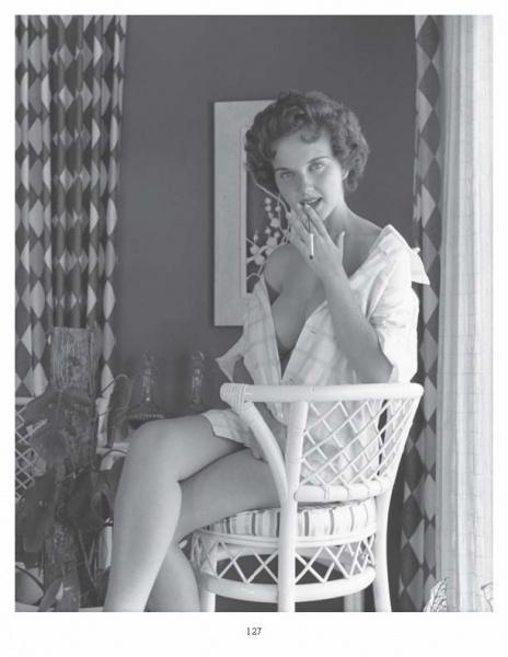 A woman sits on a wicker chair in classic 1950s style, smoking and gazing at the camera. This black-and-white image captures the allure featured in "Femmes Fatales of the 1950s" by Schiffer Publishing, Ltd. Patterned curtains and art set the scene.