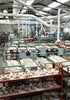 A large factory room filled with unassembled shoe parts, where Schiffer Publishing, Ltd.'s Leather Care Compendium is essential. Pipes line the high ceiling, and a worker stands near shelves in the back.