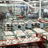 A large factory room filled with unassembled shoe parts, where Schiffer Publishing, Ltd.'s Leather Care Compendium is essential. Pipes line the high ceiling, and a worker stands near shelves in the back.