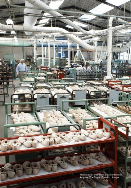 A large factory room filled with unassembled shoe parts, where Schiffer Publishing, Ltd.'s Leather Care Compendium is essential. Pipes line the high ceiling, and a worker stands near shelves in the back.