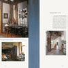 A book spread from "Passion for Primitives" by Schiffer Publishing, Ltd. shows rustic, sunlit interiors with wooden beams; the left page features a dining area and fireplace, the right a hallway and text about minimalist decor.