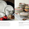 Two catalog pages: The left features white braided-rim ceramic dinnerware with a mug, tomatoes, and bread. The right displays vibrant, patterned serving dishes, echoing the handcrafted style showcased in Celebrating MacKenzie-Childs by Schiffer Publishing, Ltd.