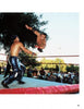 A wrestler soars toward an opponent in an outdoor ring as a referee and crowd watch under sunlit trees—a thrilling scene from Hard Boys and Bad Girls by Schiffer Publishing, Ltd., inspiring aspiring wrestlers everywhere.