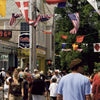 A lively street scene in Provincetown, MA, with people strolling beneath colorful flags and a sign reading 96°F. Shops and trees line the sunlit avenue—just one of many captivating images in "Provincetown Perspectives" by Schiffer Publishing, Ltd.