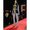 A model presents the latest look from "Emerging Fashion Designers 2" by Schiffer Publishing, Ltd., wearing a sleeveless navy vest with yellow accents and grey pants on a red runway as spectators watch, with a large letter E seen in the background.